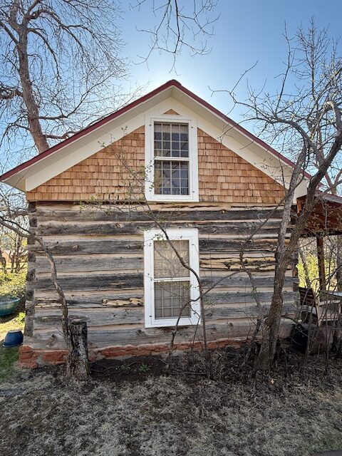 A weathered hand-hewn log cabin with cedar-shingle gable and white-trimmed windows